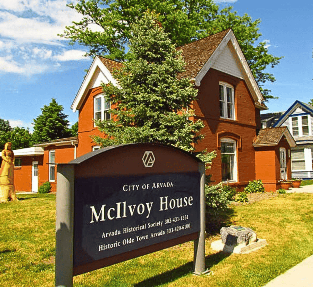 Historic McIlvoy House in Arvada, Colorado, part of the Olde Town historic district, with lush greenery and a clear blue sky.