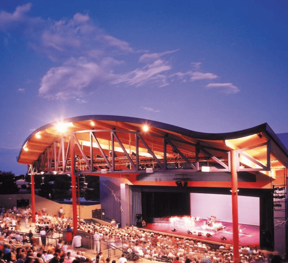Vibrant outdoor theater at dusk with an illuminated stage and an audience enjoying a live performance.