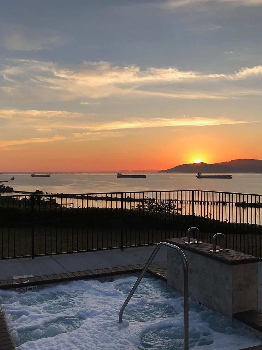 Breathtaking sunset view over the water from a hot tub, with ships sailing and a scenic horizon in the background.