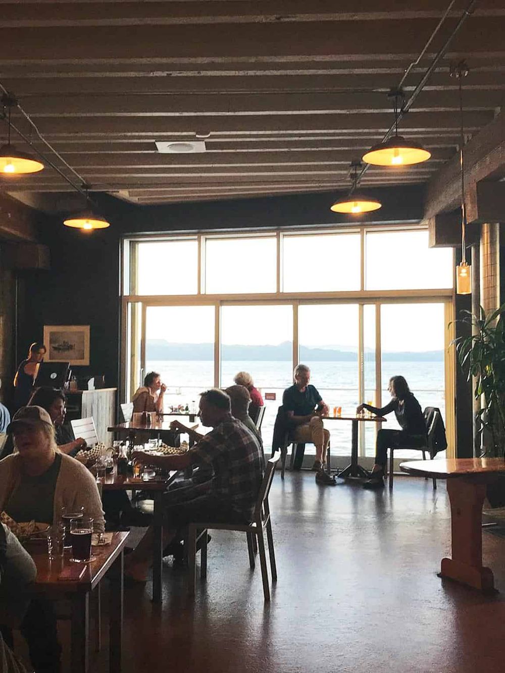 A cozy waterfront restaurant with large windows overlooking a lake, featuring warm lighting and guests enjoying their meals.
