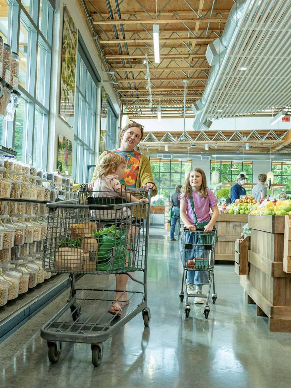 Bright indoor farmer's market with families shopping for fresh produce and groceries, featuring large windows and modern wooden interior.