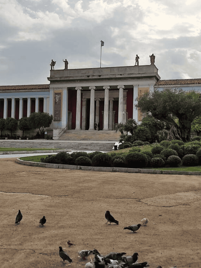 Ancient Greek-style building with statues and pigeons in front, on cloudy day.