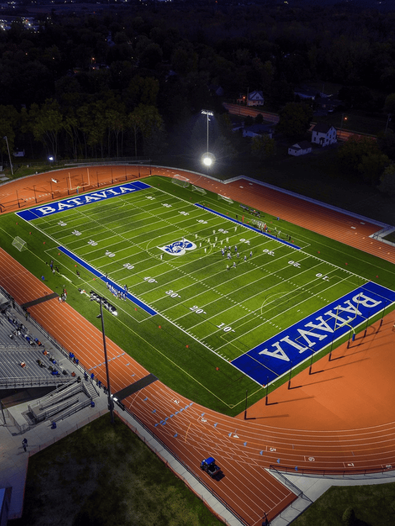 Brightly lit high school football field at night with track surrounding, marked with "BAYLA" and football logo.
