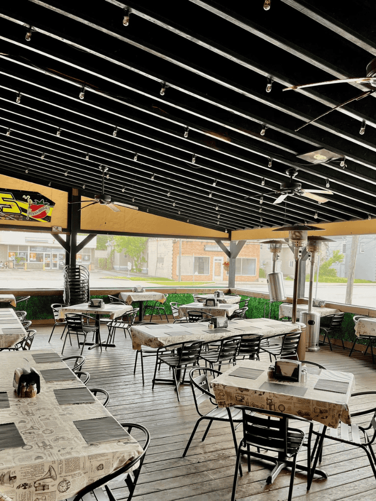 Outdoor restaurant dining area with tables, chairs, and heaters on a wooden patio.
