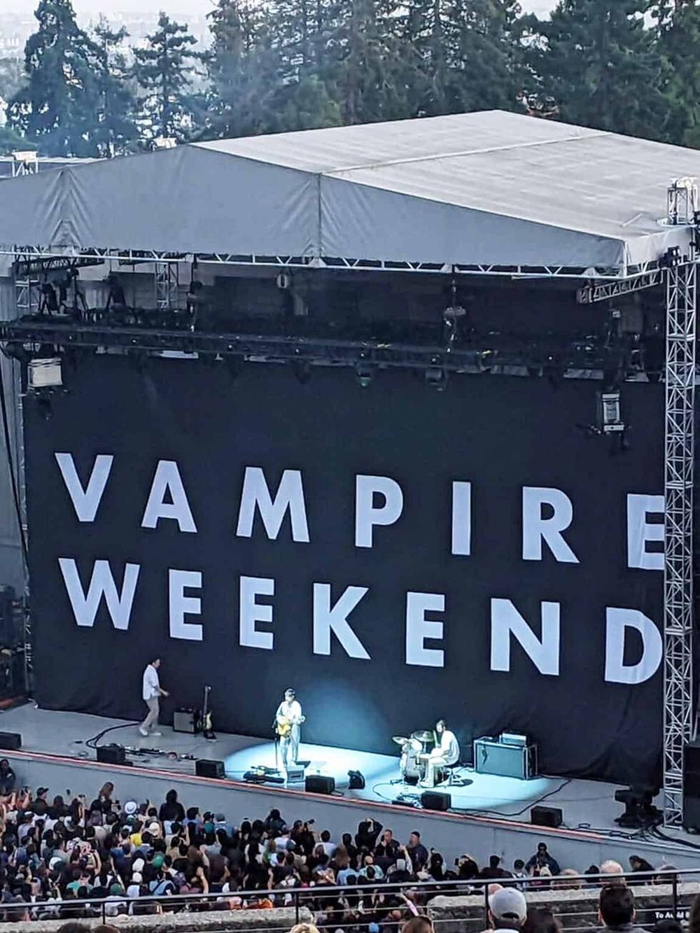 Vampire Weekend live concert stage with large black backdrop and audience in outdoor setting.