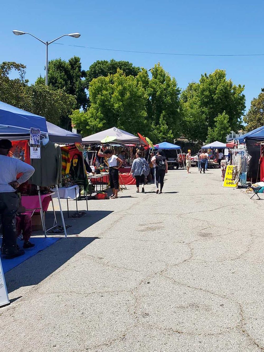 Stalls at an outdoor market, with trees and vendor tents under a clear blue sky, showcasing local shopping and community events.