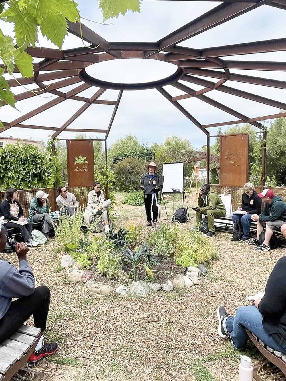 Outdoor garden workshop or seminar with people seated around a circular garden bed, led by a speaker under a unique open structure.