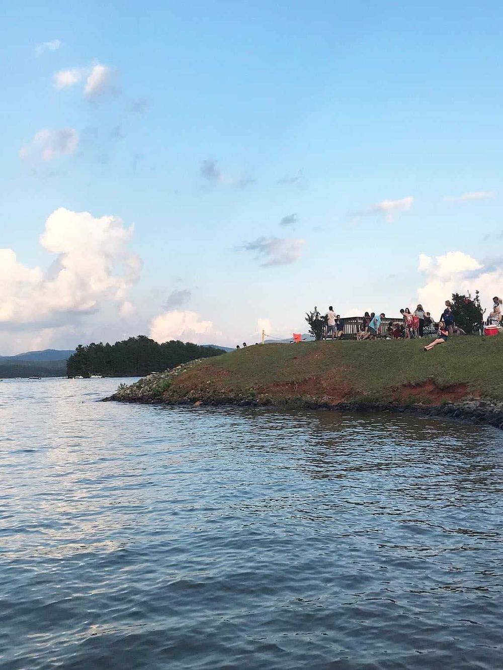 People enjoying a scenic lakeside view with a grassy hill and a observation deck during daytime.