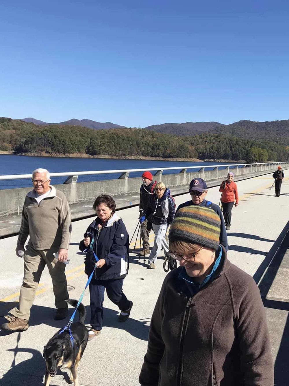 Group of people walking on a bridge by a lake with mountains in the background, enjoying outdoor adventure and nature.