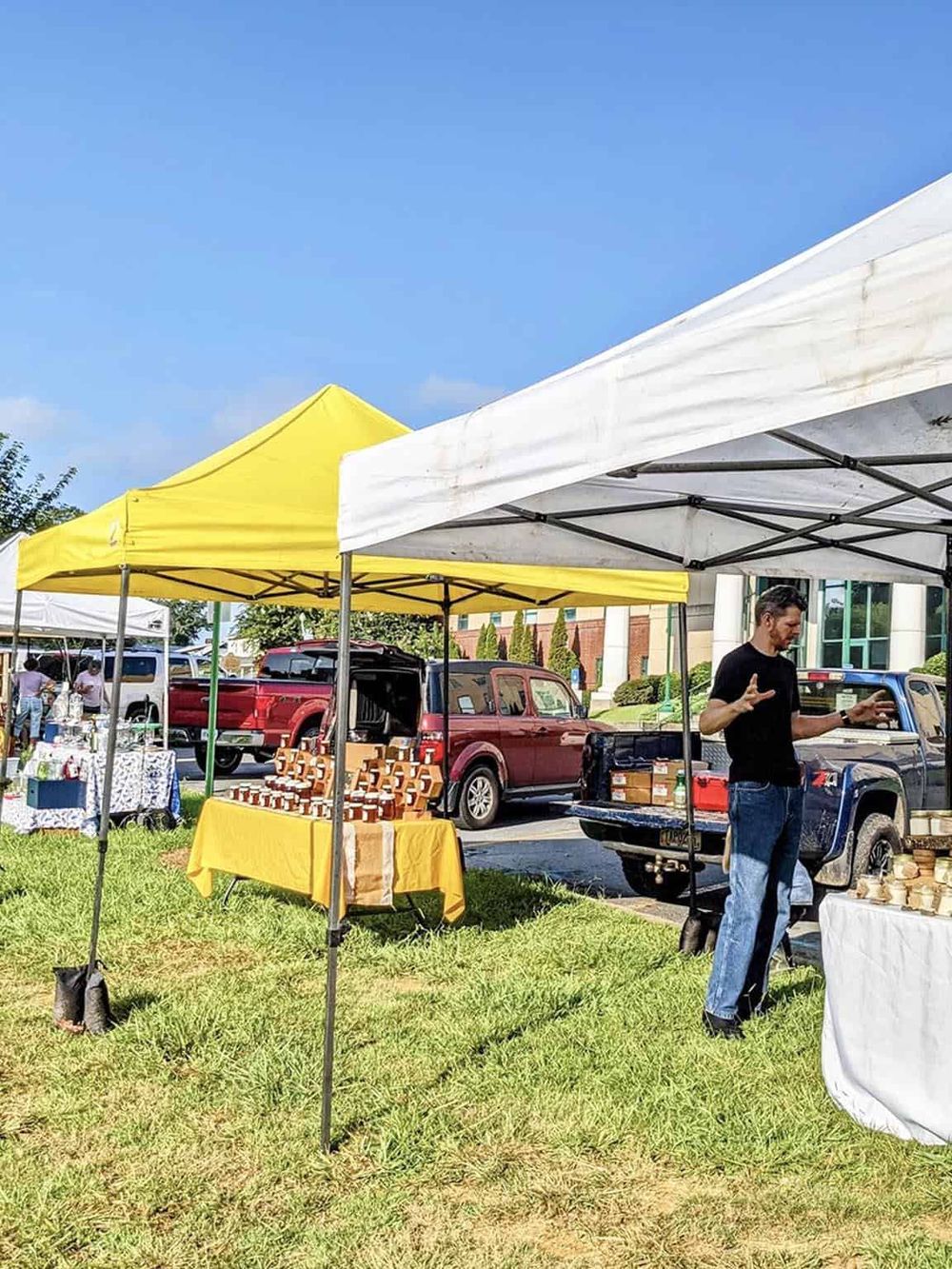 Colorful outdoor farmers market stalls with vendors and vehicles on a sunny day.