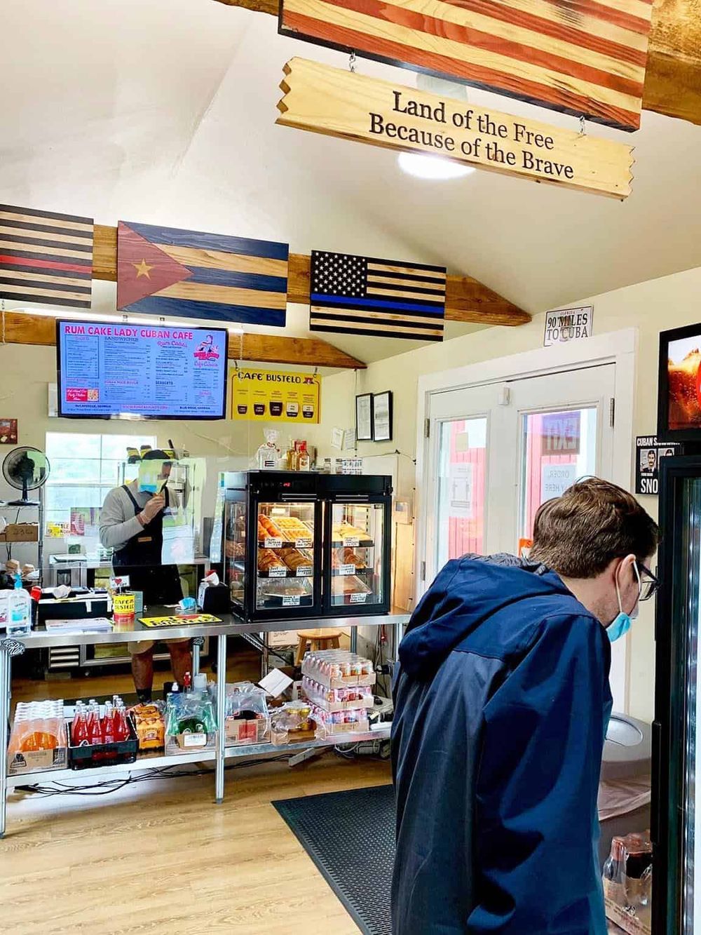Cuban café with bakery display and customers ordering, vibrant atmosphere, tropical décor, and flags.