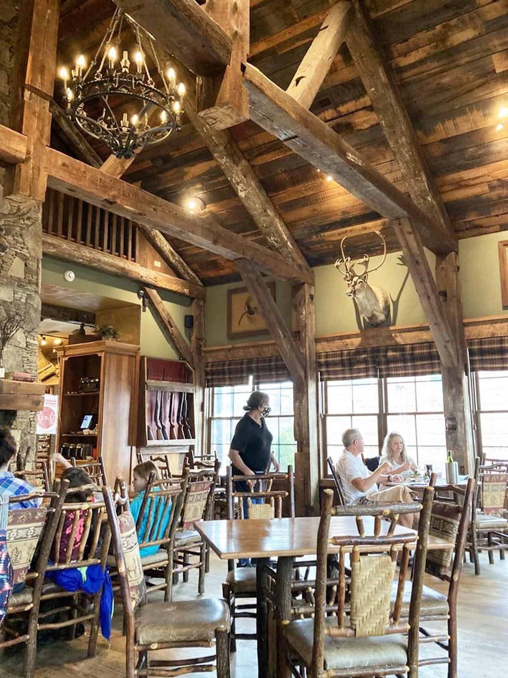 Rustic lodge restaurant interior with wooden beams, chandeliers, and a mounted deer head. Family dining in cozy mountain retreat ambiance.