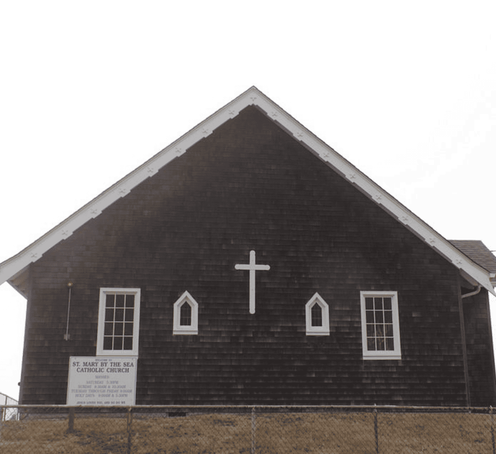 Old wooden church building with cross and sign, historical site of St. Mary by the Sea Catholic Church.