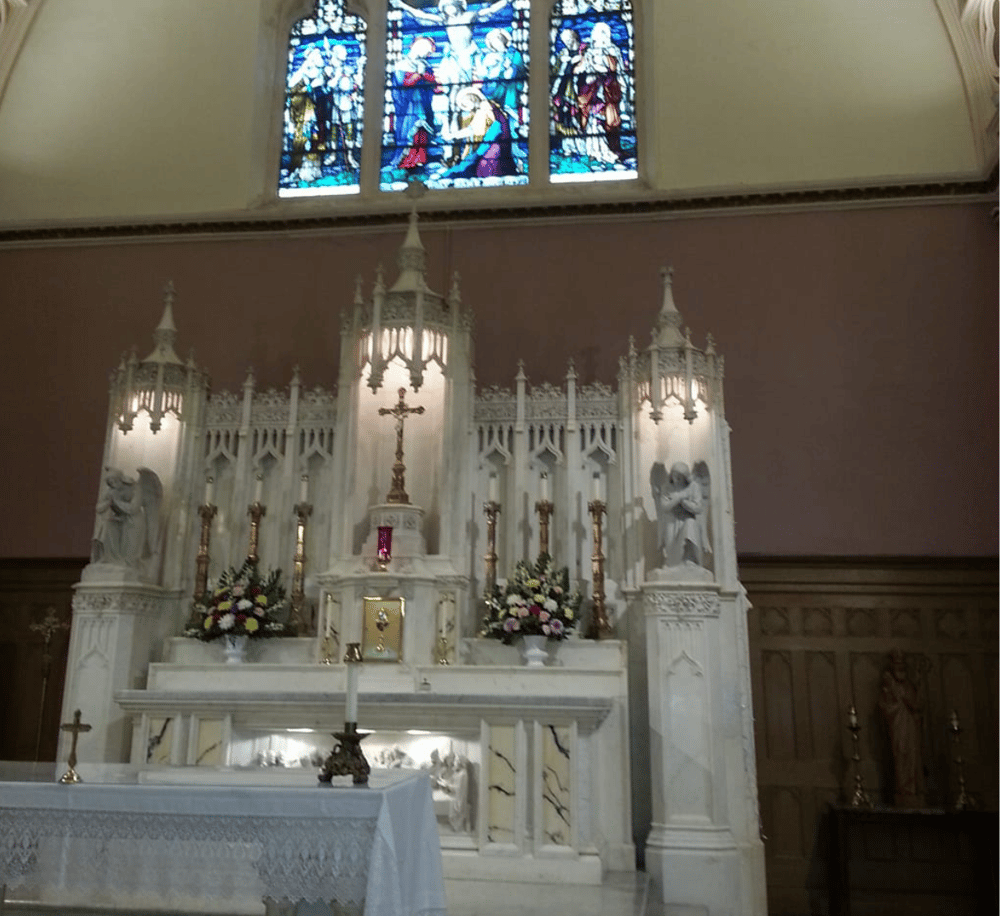 Ornate church altar with stained glass window, religious statues, and floral arrangements.