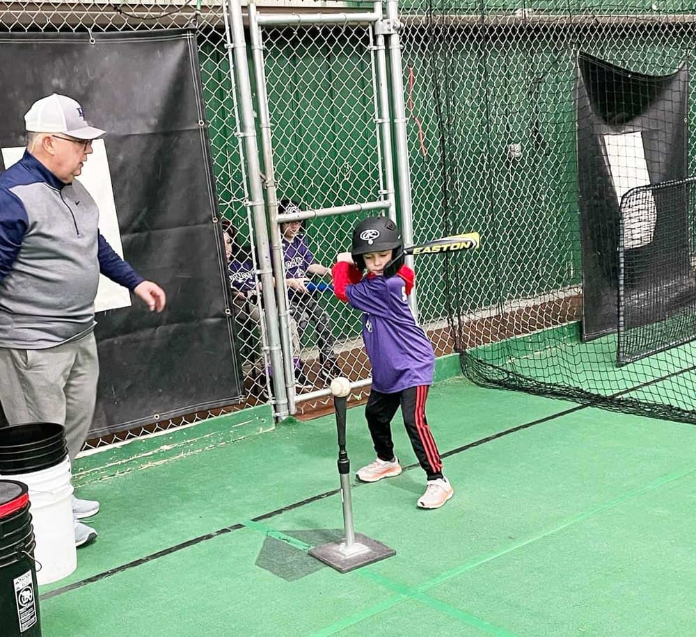 Young baseball player practicing batting with coach on a batting tee at a training facility.