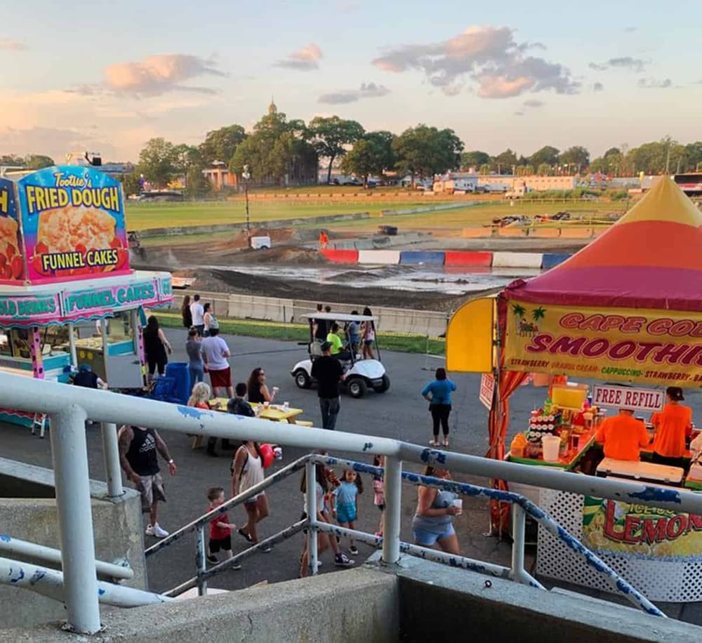Colorful fairground with food stalls, amusement rides, and a dirt race track at sunset.