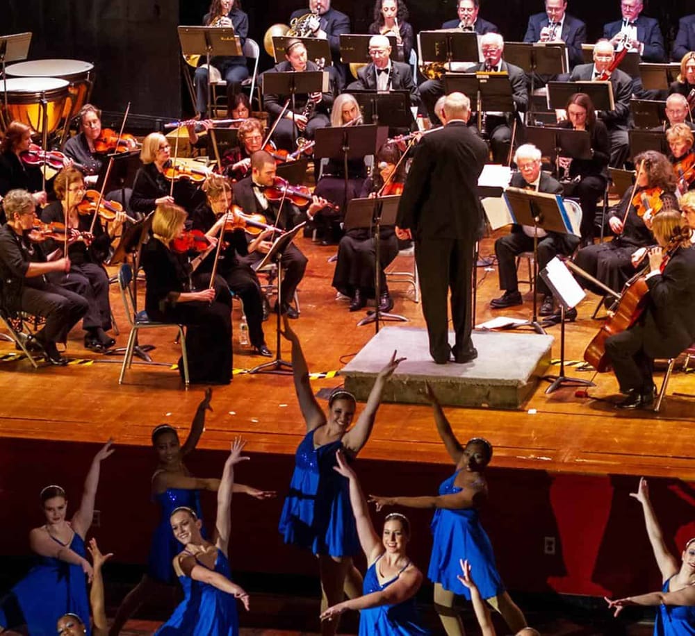 Orchestra performing with ballet dancers in blue costumes on stage, showcasing live music and dance entertainment.