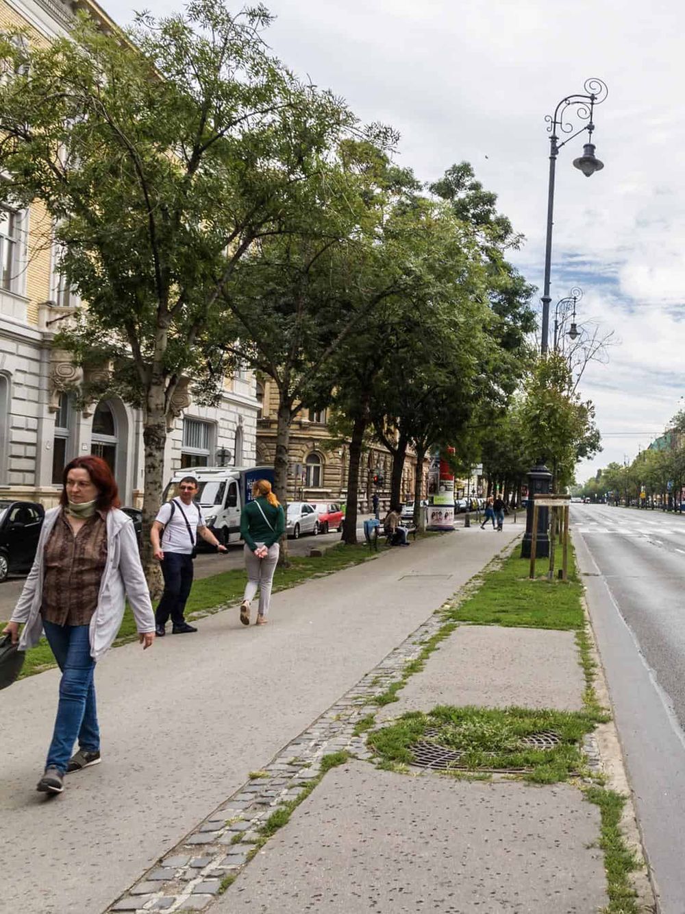 Broad city street sidewalk with pedestrians, trees, vintage lampposts, and urban architecture.