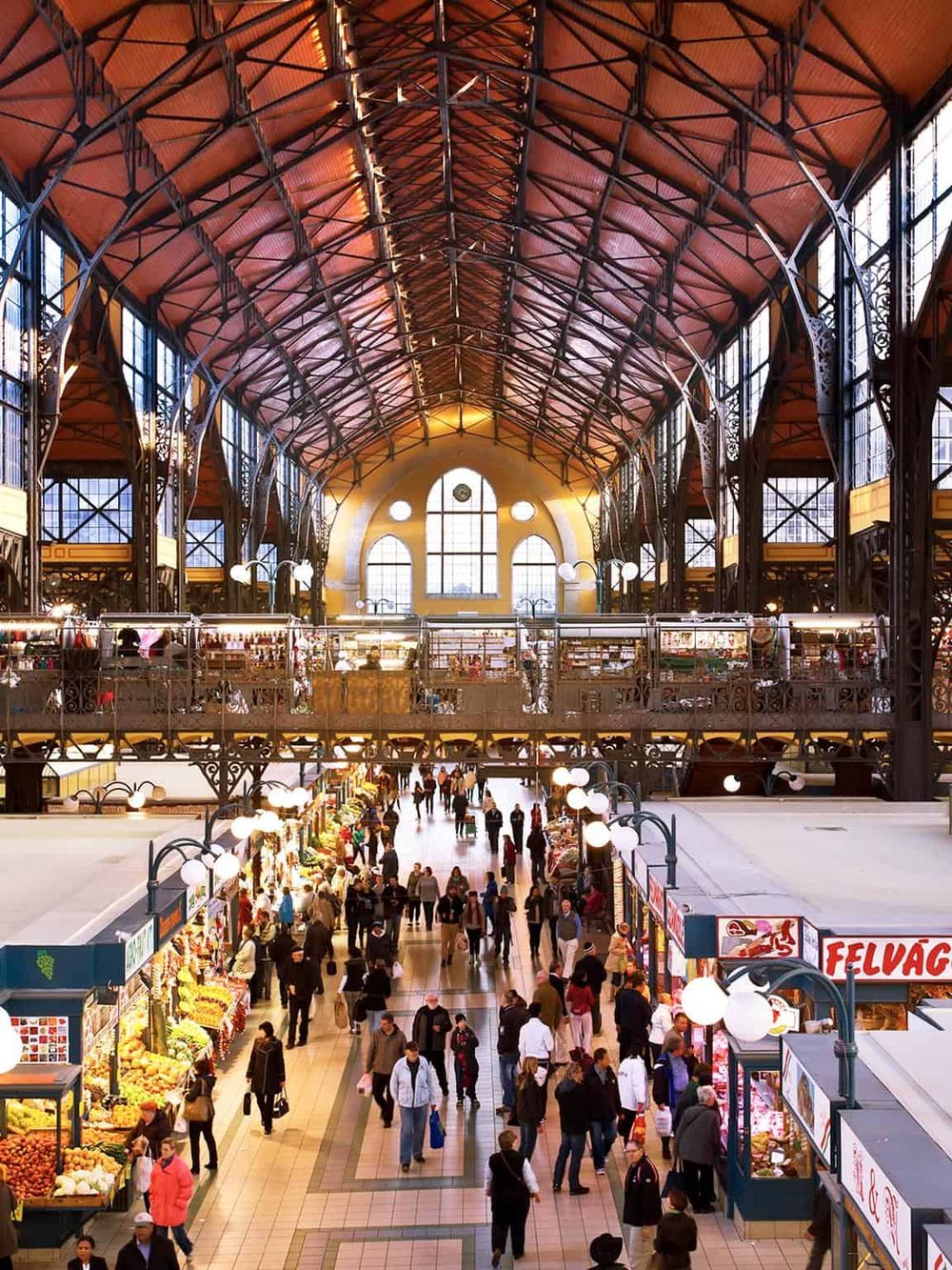 Vibrant indoor marketplace with natural light, bustling shoppers, and food stalls under a historic iron-and-glass ceiling.