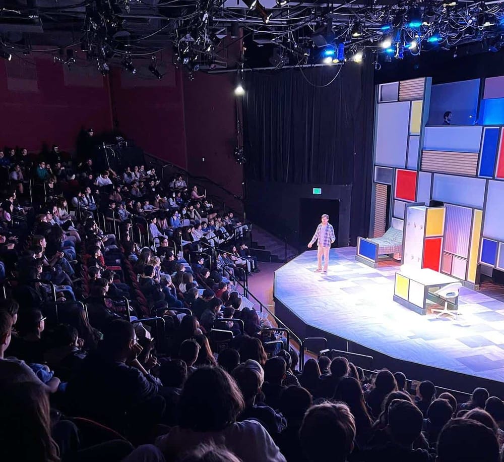 A vibrant theater stage with colorful geometric set design hosting a live performance, audience seated in a darkened auditorium for a comedy show.