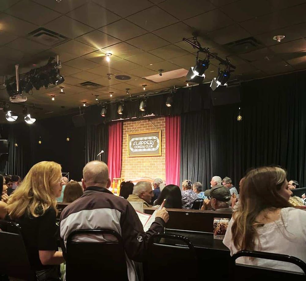 Adult comedy club audience seating scene with stage, lighting, and brick backdrop.