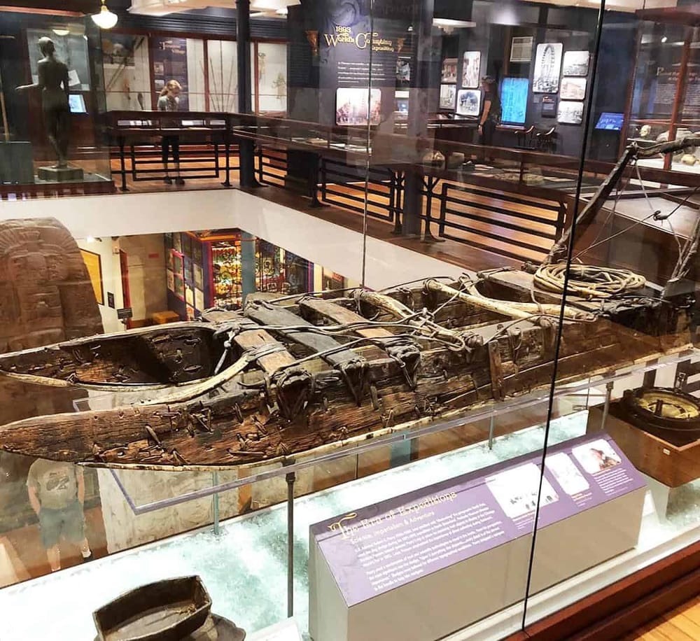 An ancient wooden boat exhibit displayed in a museum's glass case with museum visitors in the background.