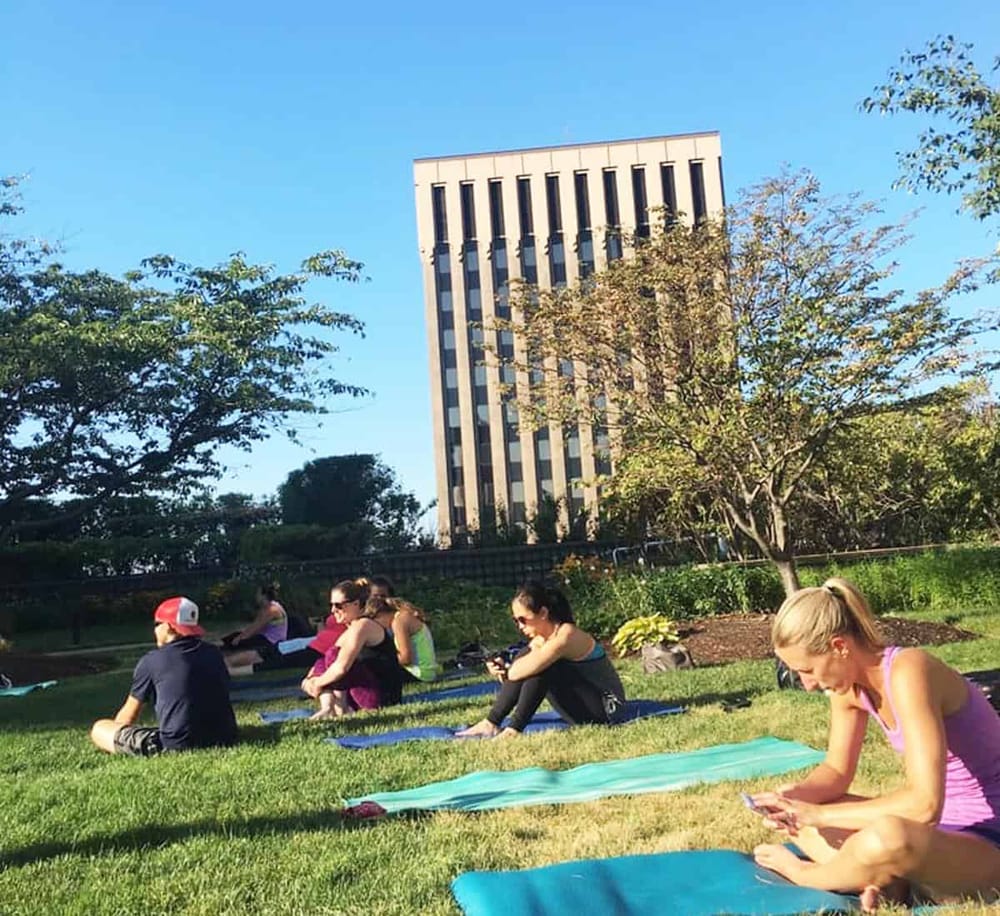 Relaxing outdoor yoga class in urban park with city skyscraper background, sunny weather, and peaceful atmosphere.
