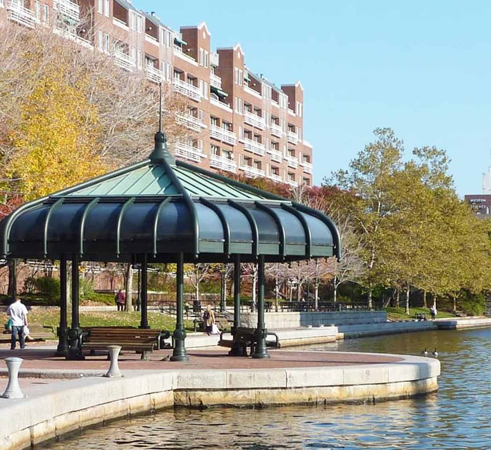 Urban park gazebo overlooking water, city skyline with residential buildings in the background.
