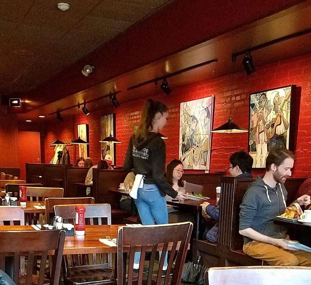 Cozy restaurant interior with guests dining and an attentive server, vibrant artwork on brick walls enhancing the ambiance.