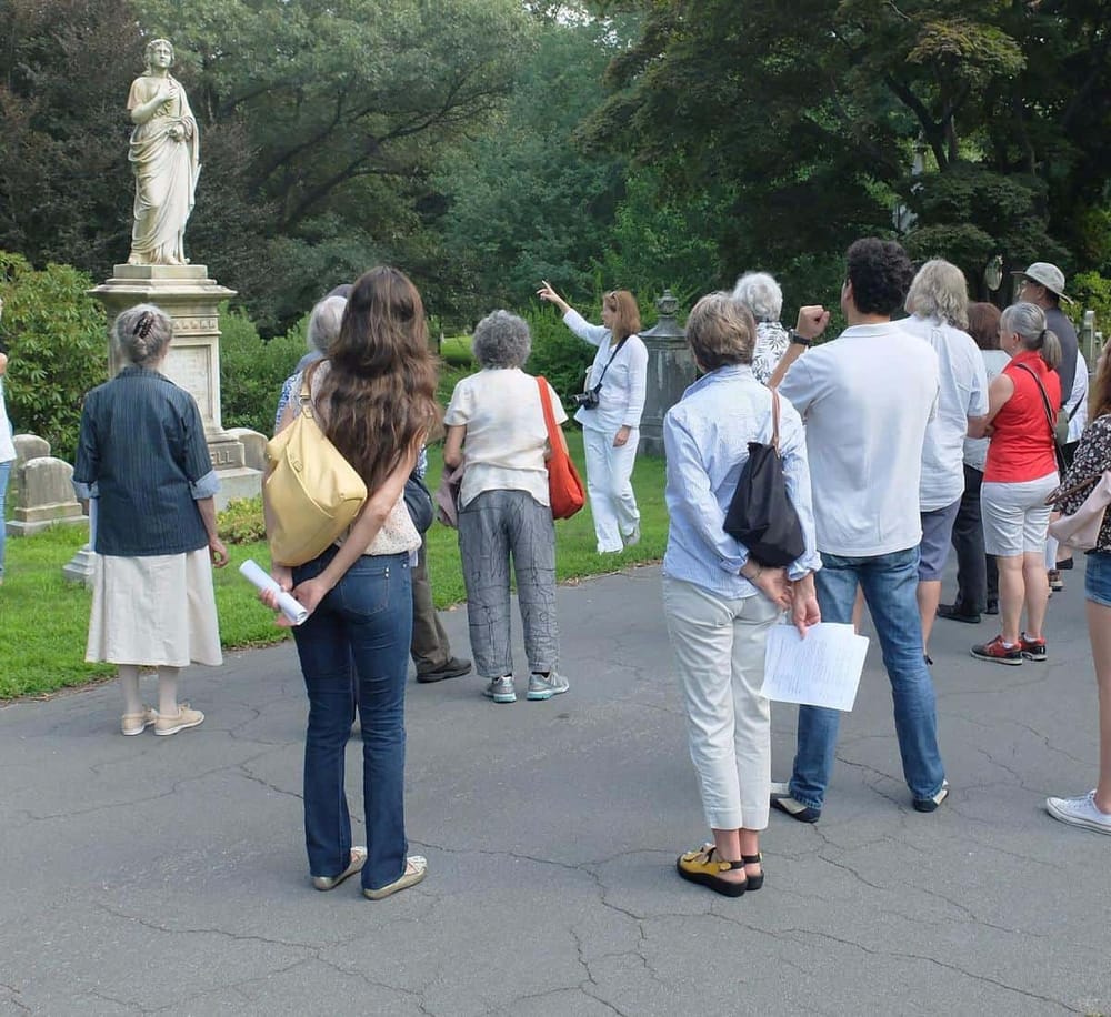 Historic cemetery tour guides and visitors exploring graves and statues in lush green park setting.