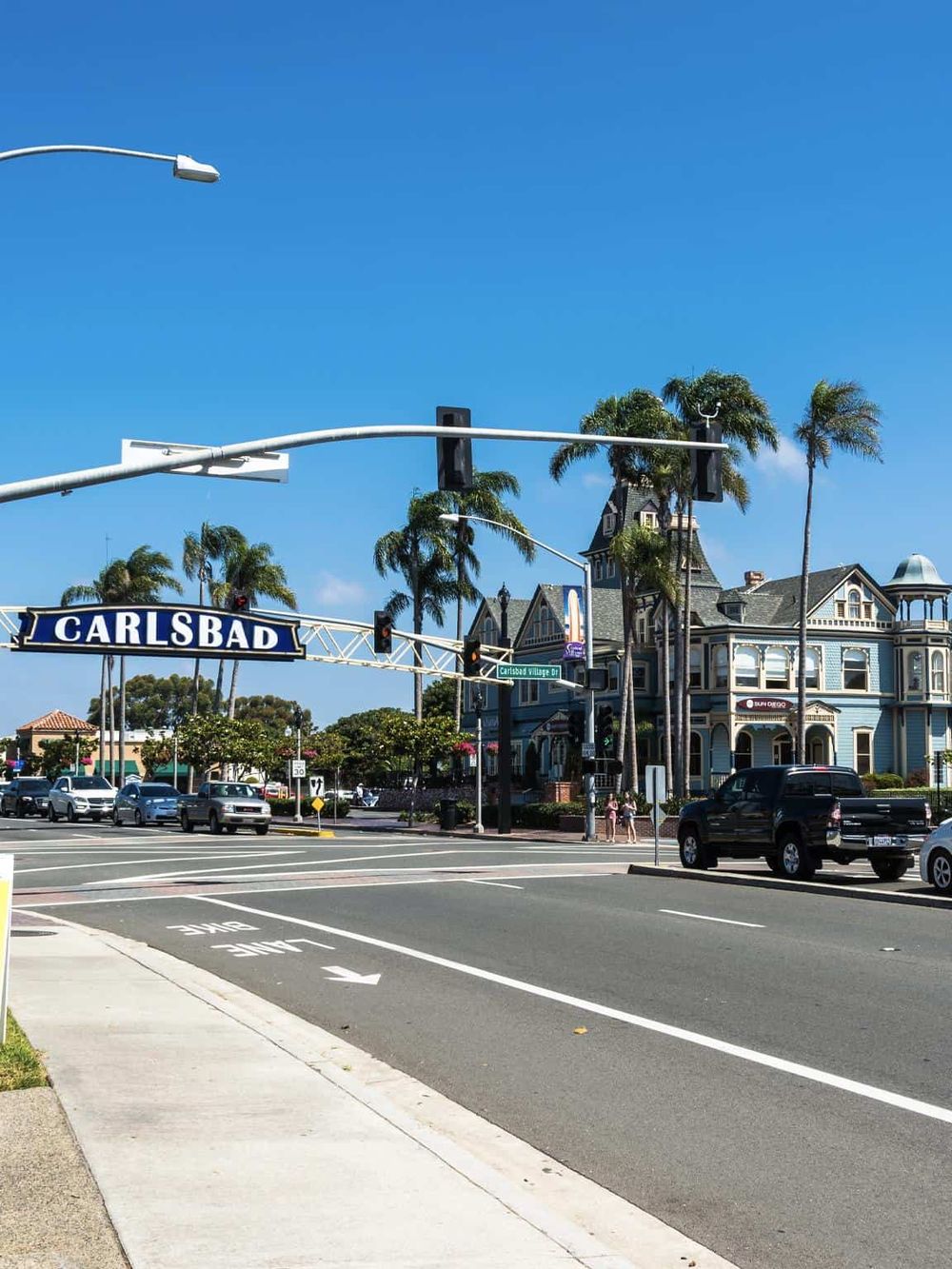 Colorful Victorian-style building with palm trees on a sunny day in Carlsbad, California, featuring roadside traffic.