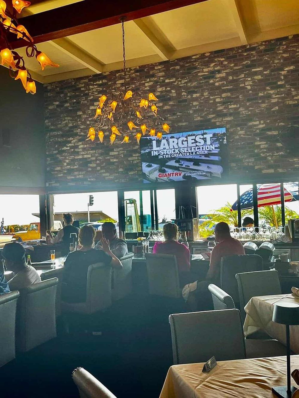 Elegant restaurant interior with large TV displaying stock news, hanging floral chandelier, and panoramic views through windows.