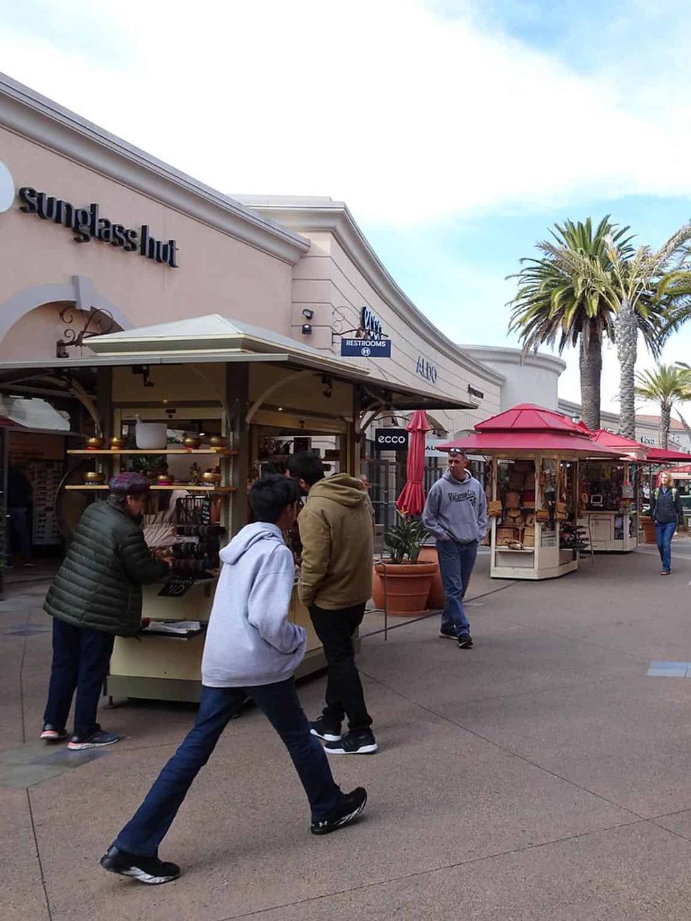 Vibrant shopping district with retail shops and outdoor kiosks, palm trees, and shoppers at Quest for Directions.