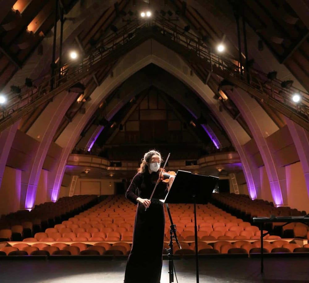 Elegant woman playing violin in concert hall with wooden architecture and stage lighting.