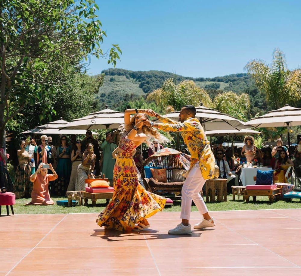 Colorful outdoor wedding celebration with dancing and guests enjoying sunny day in scenic garden setting.