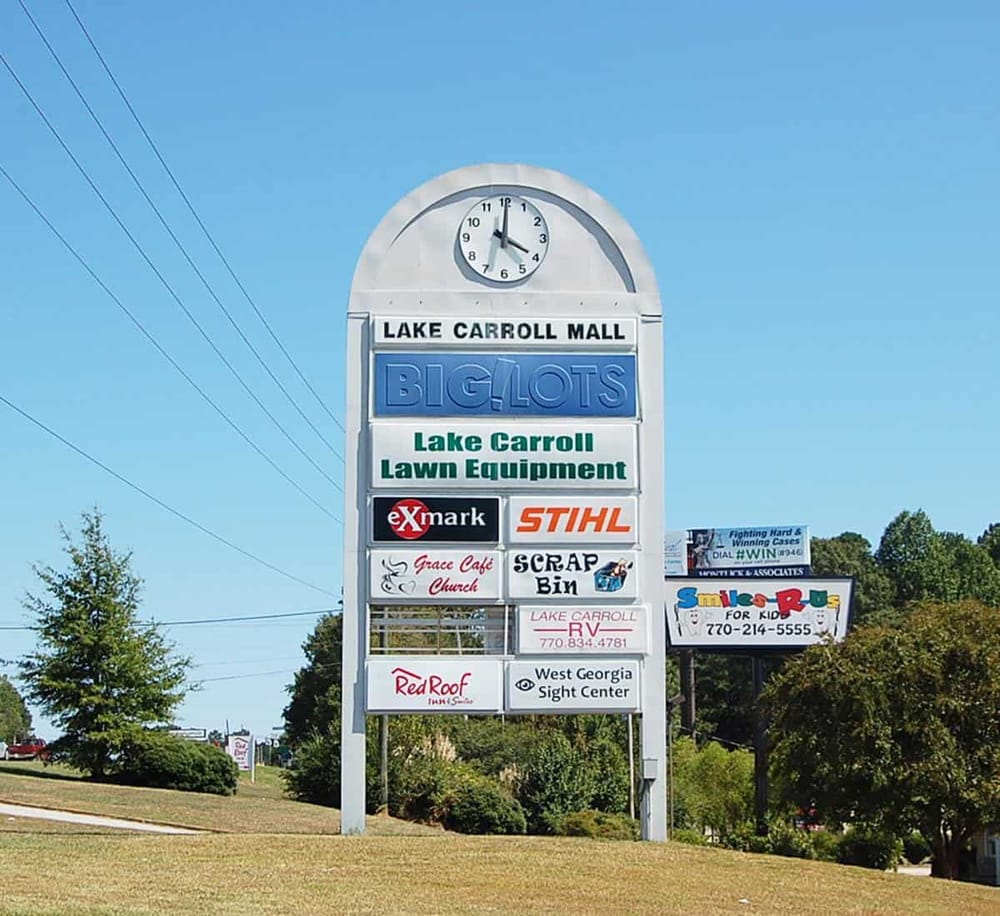 Large outdoor shopping mall sign with store names in Lake Carroll, Georgia, with clear sky.
