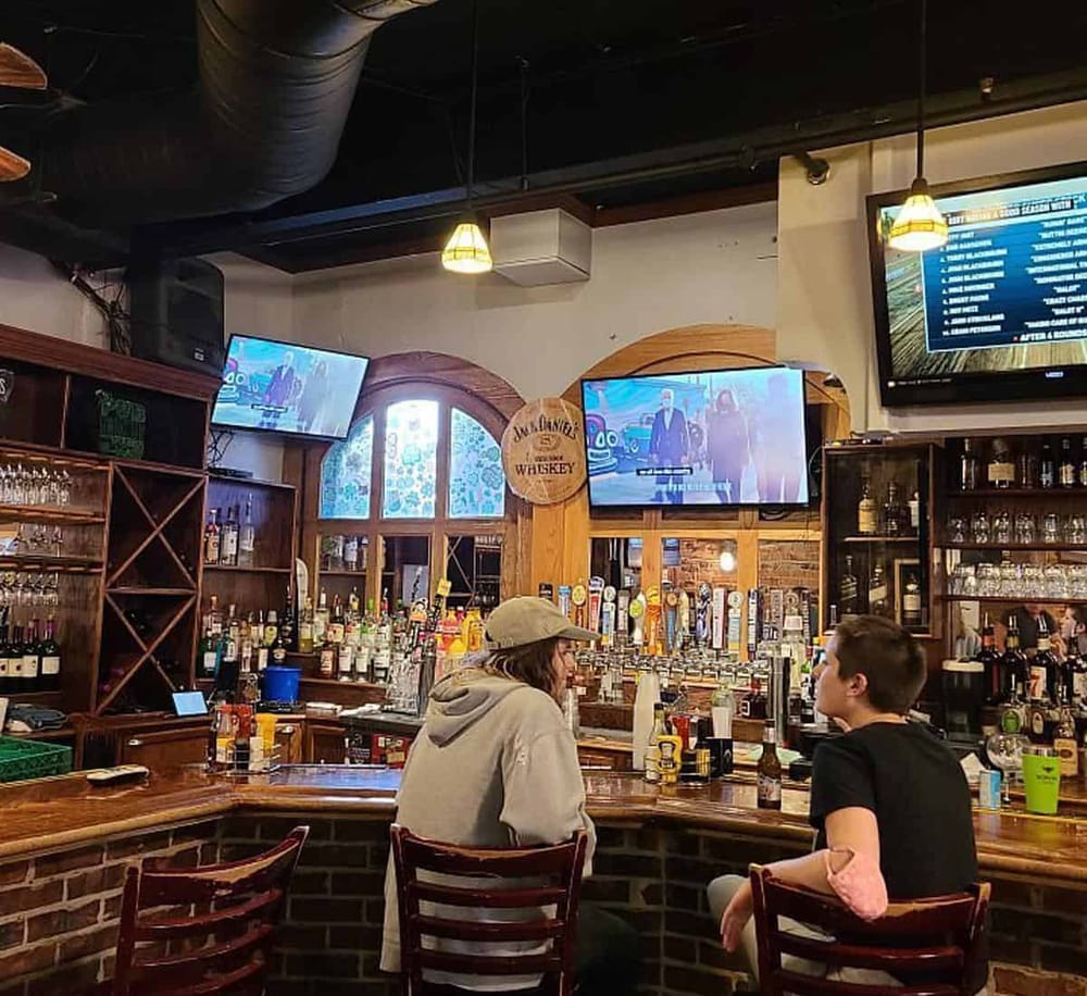 Cozy bar scene with two patrons talking, multiple TV screens, and a well-stocked alcohol display.
