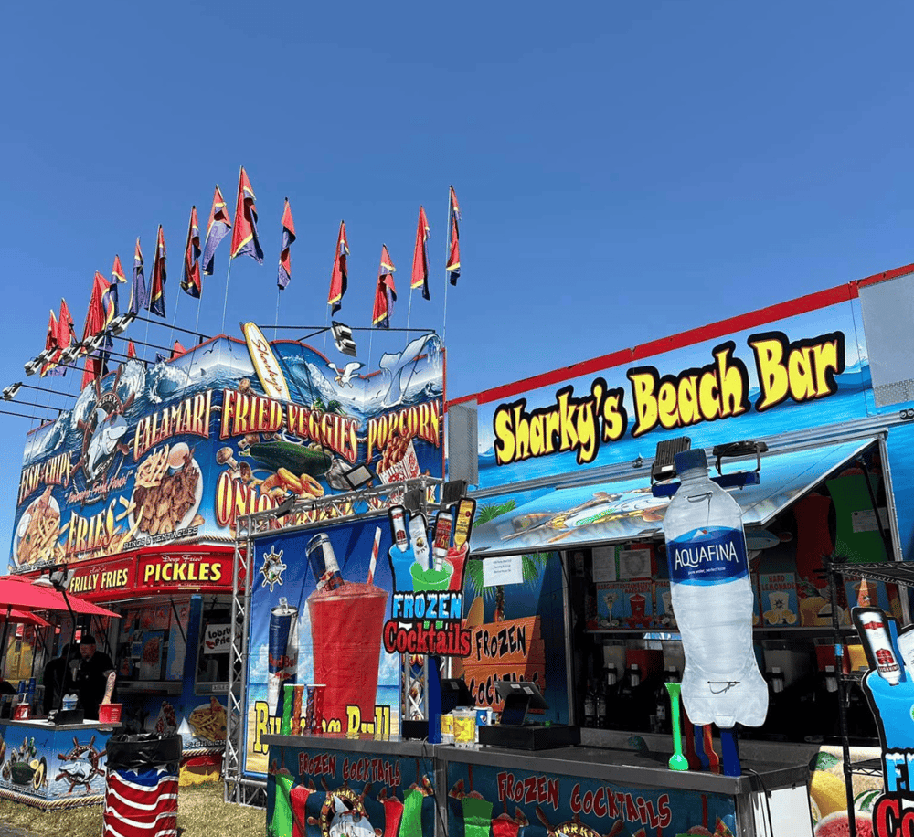 Colorful seaside food stand at a beach fair with drinks and snacks, vibrant signs, and flags under a bright blue sky.