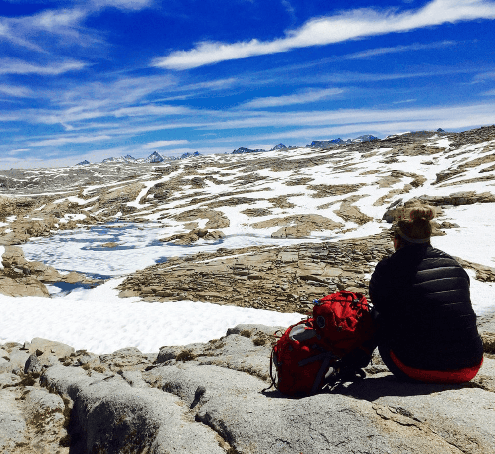 Scenic mountain hiking landscape with snow, rugged terrain, and a hiker resting with a backpack.