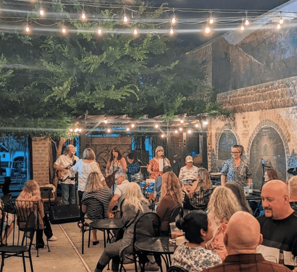 Live outdoor evening band performance at QuestForDirections venue, featuring string lights, rustic decor, and social ambiance.