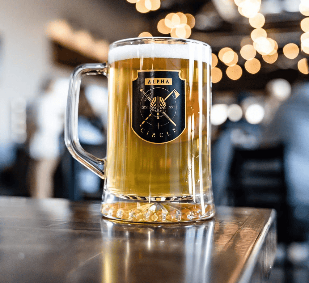 Cold beer in a clear glass beer mug with black and gold "Alpha Circles" logo, on a wooden table at a bar or pub.