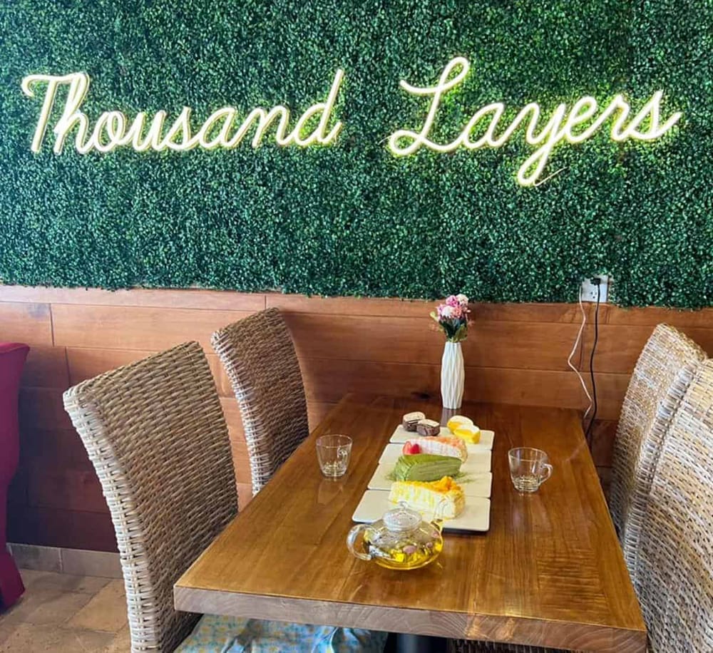 Neon "Thousand Lays" sign on green leafy wall with cozy dining setup and desserts on the table.