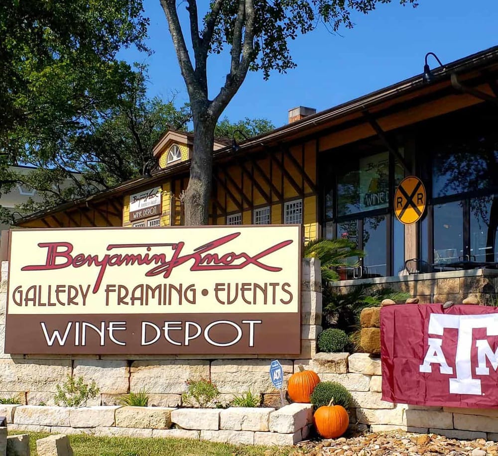 Colorful Benjamin Knox gallery and wine depot sign in front of charming yellow building with autumn pumpkins and lush trees.