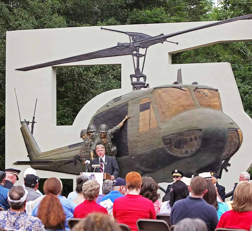 Vintage military helicopter memorial at QuestForDirections outdoor event, with speaker and audience attending.
