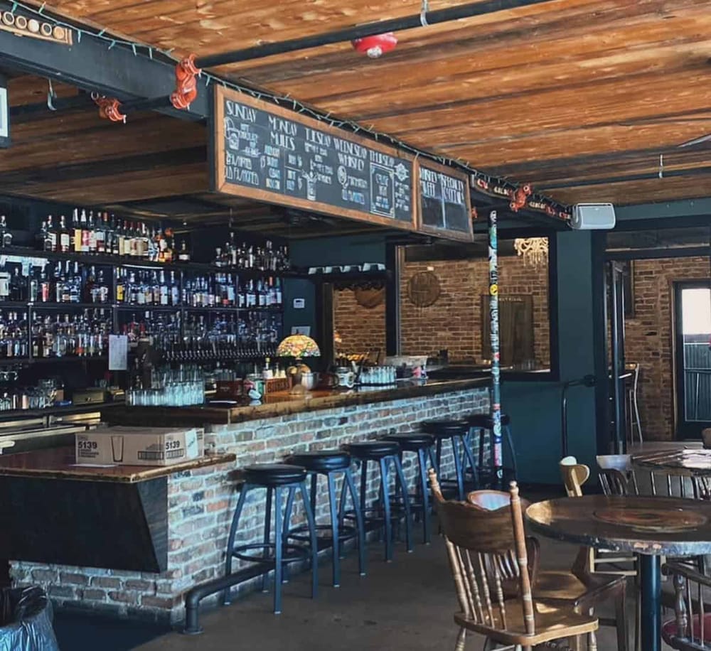 A cozy bar interior with exposed brick walls, wooden beams, and a fully stocked liquor shelf. Perfect for socializing and drinks.