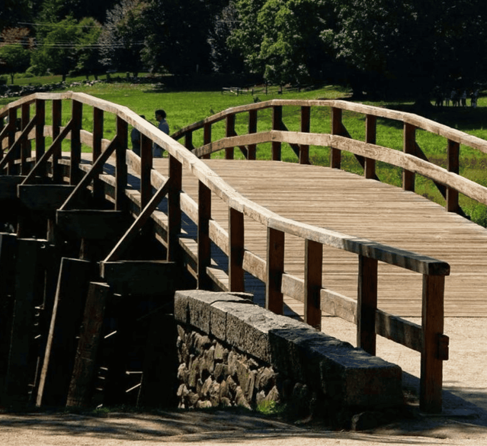 A rustic wooden pedestrian bridge in a park setting with lush green grass and trees.