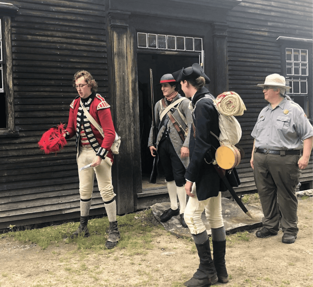 Colonial reenactors in historical costumes outside a wooden building for educational demonstration.