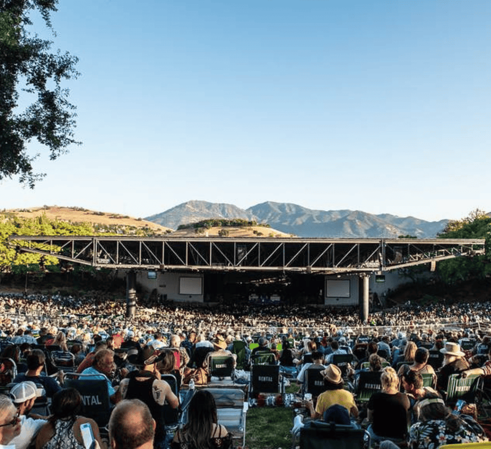Crowd at outdoor concert with stage, mountain backdrop, and evening sky.