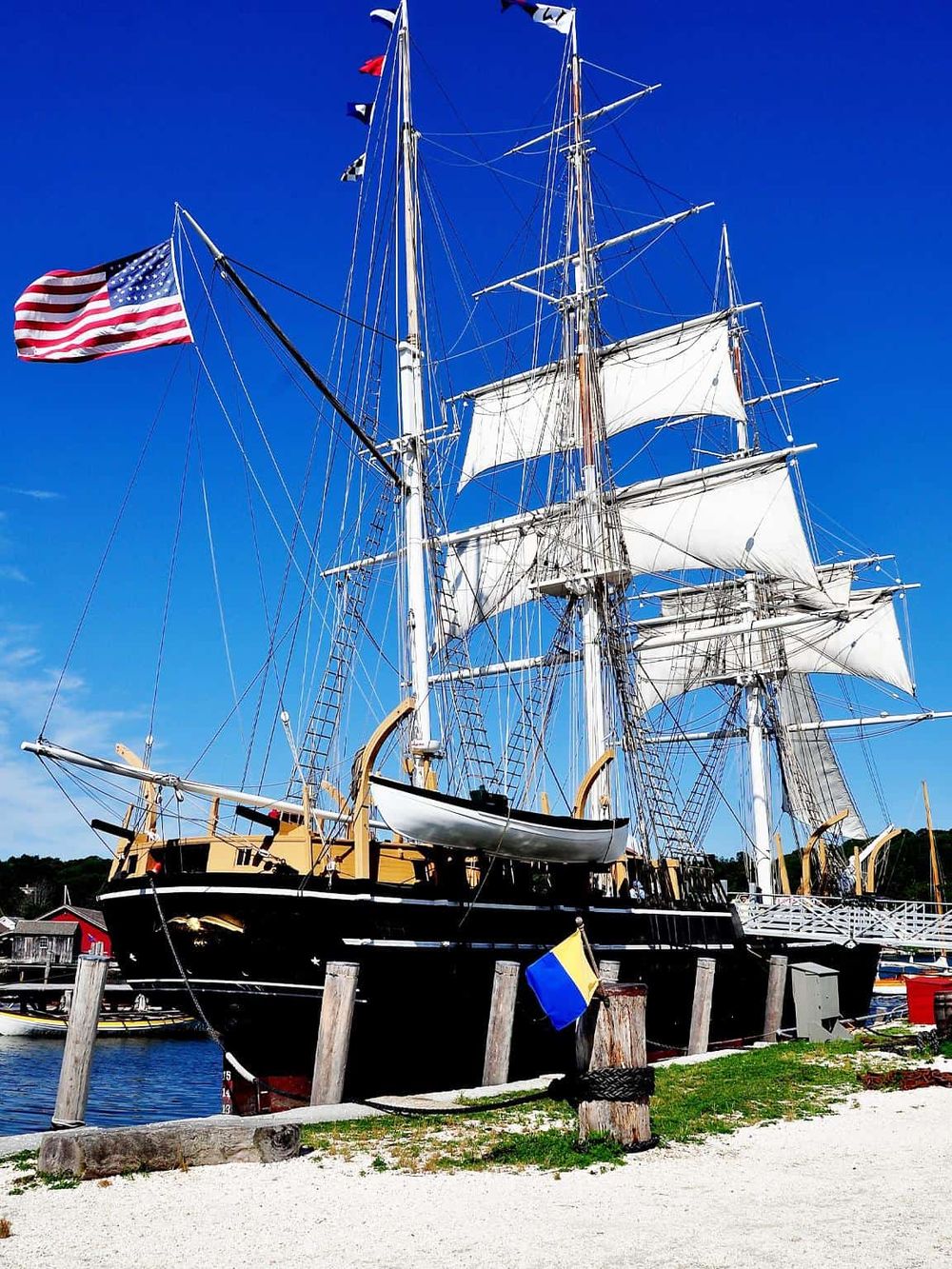 Majestic sailing ship docked at harbor with American flags, picturesque blue sky, and waterfront scenery.