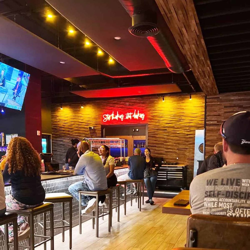 Modern bar interior with people socializing and beverages, stylish wood accents, and vibrant neon signage.